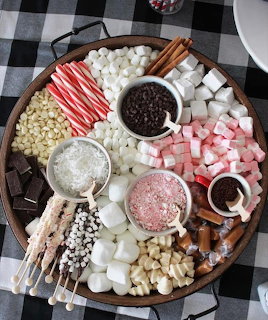 Round wooden board with marshmallows, candy canes, chocolate chips, and hot cocoa toppings on plaid cloth
