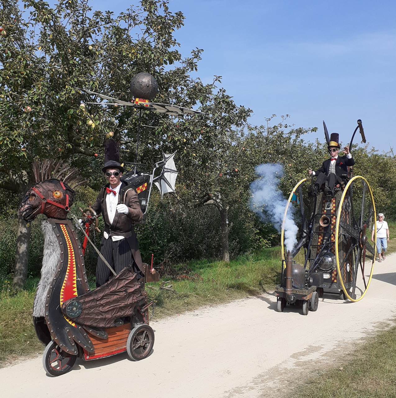 Two people in steampunk outfits ride whimsical mechanical machines on a tree-lined dirt path.