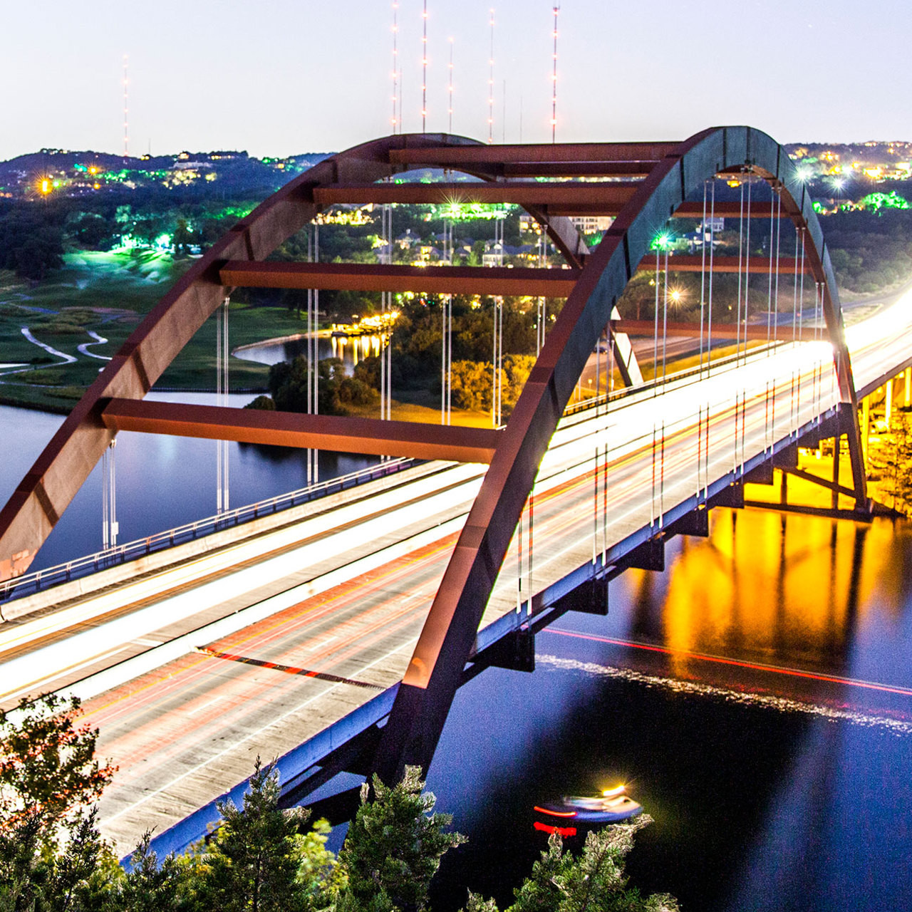 Red arch bridge lit at night with light trails crossing over a river, city lights in background