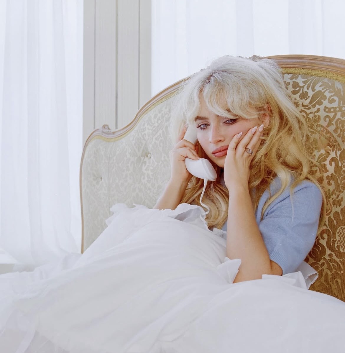 Blonde woman in blue outfit reclining on ornate cream and gold vintage bed