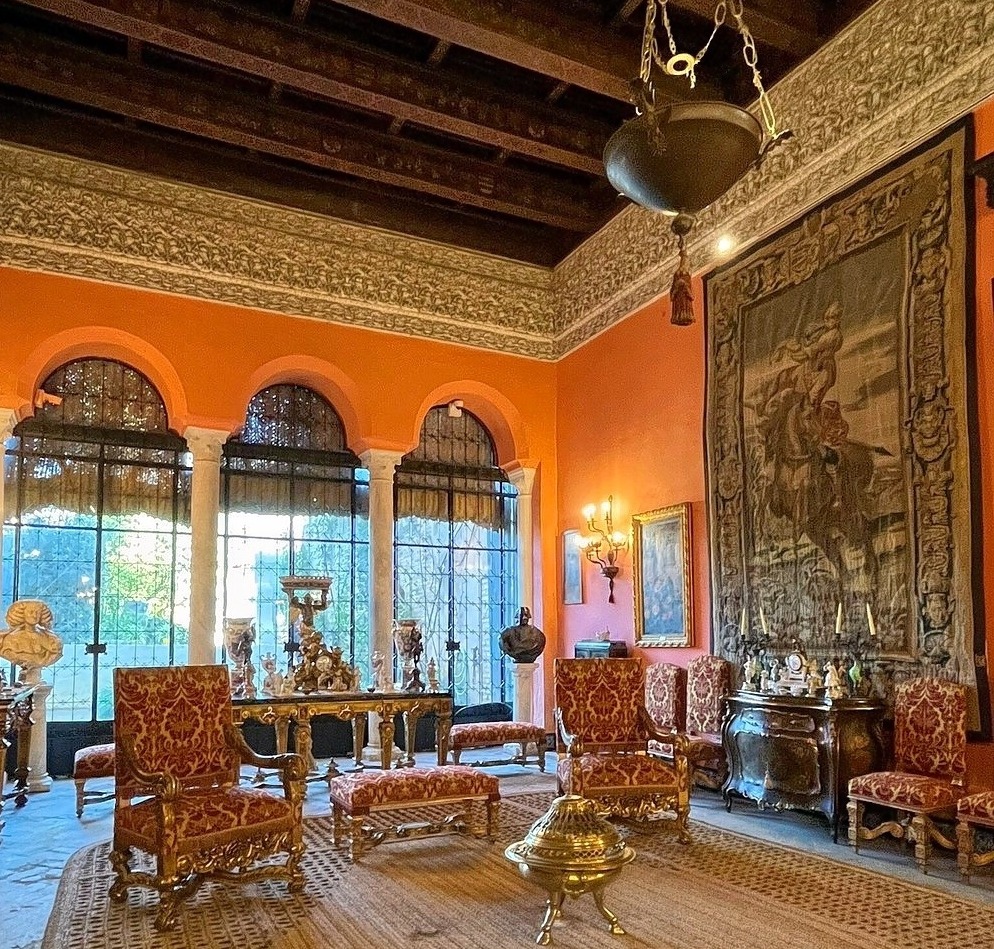 Ornate palace room with orange walls, arched windows, antique chairs, and a large wall tapestry