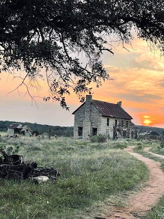 Small stone cottage by a dirt path in a grassy field at sunset with tree branches overhead