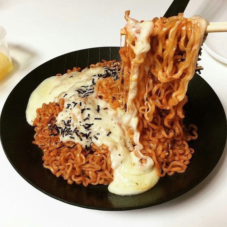 Chopsticks lifting spicy orange ramen noodles with melted cheese and sesame seeds from a black bowl