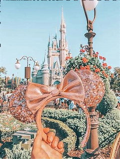 Hand holding sparkly rose gold Mickey ears with bow in front of Disney castle and topiaries