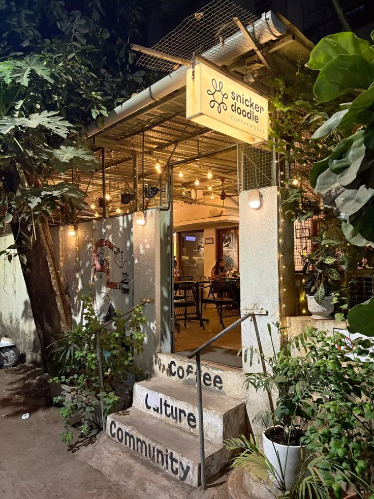 Illuminated coffee shop entrance at night with tropical plants and wooden structure