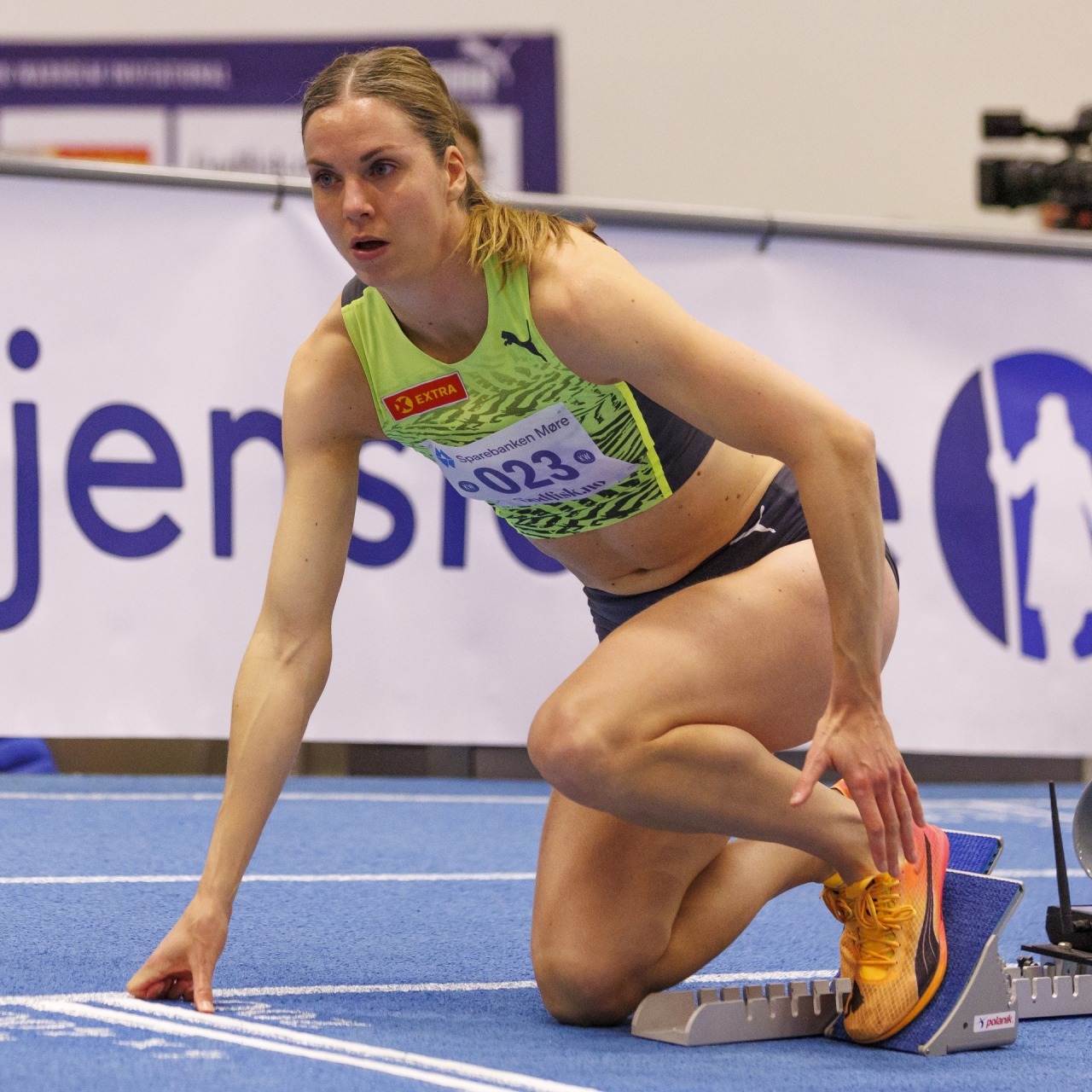 Female athlete in yellow top crouched in starting blocks on blue indoor track ready to race