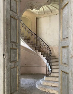 Spiral stone staircase with decorative iron railing viewed through open wooden doors in aged Italian building