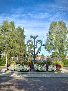 Metal sculpture with bird and flower motifs in park garden with white blooms and birch trees