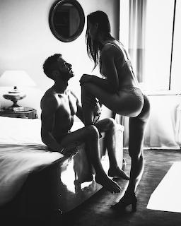 Black and white photo of a couple in intimate pose in a bedroom, woman in lingerie standing near seated man