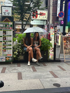 Two women sitting under a clear umbrella on a rainy city sidewalk in Japan