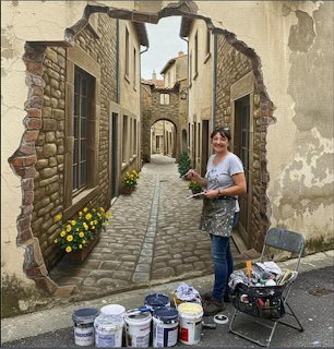 Woman artist posing next to a realistic mural of a European stone alley painted on a wall