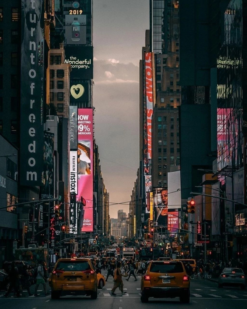Busy Times Square street at dusk with tall buildings, neon signs, yellow taxis, and pedestrians