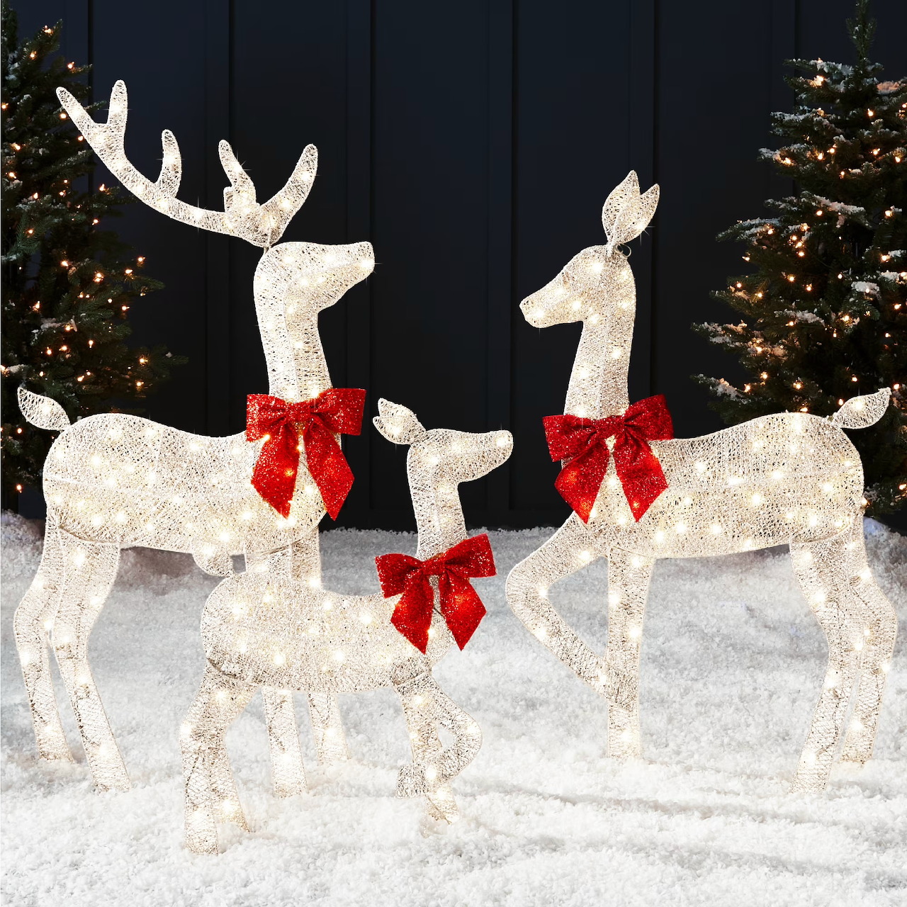 Three glowing white reindeer yard decorations with red bows in snow with Christmas trees