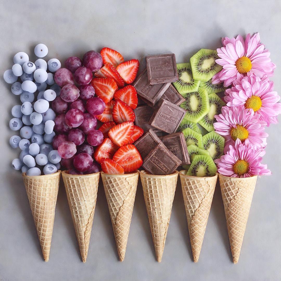 Waffle cones filled with berries, chocolate, kiwi, and flowers on gray background
