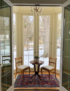 Sunroom with two wooden chairs, round table, oriental rug, and tall windows overlooking snowy yard