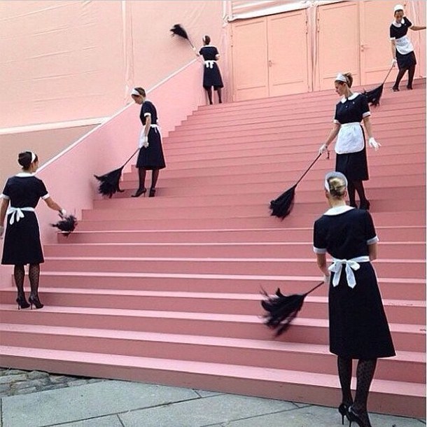 Multiple women in black and white maid uniforms holding brooms, positioned on wide pink stairs