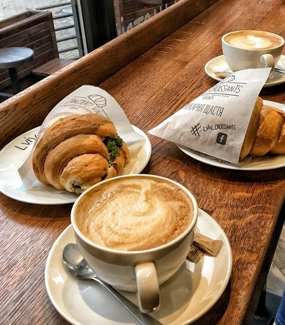 Cafe table with two cappuccinos, a croissant in paper wrapping, and a stuffed sandwich on wooden counter