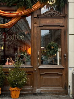 Wooden coffee shop entrance with glass door, window sign, wreaths, orange drapery, and potted plants