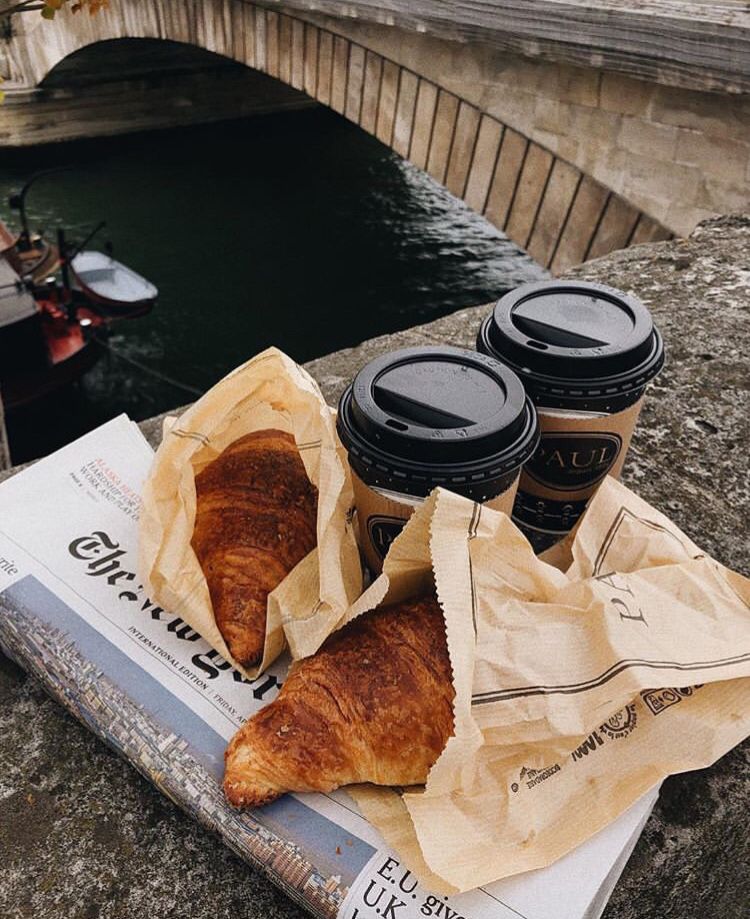Two croissants and two takeaway coffee cups on a newspaper by a canal with a stone bridge