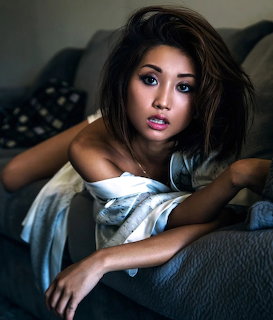 Young Asian woman with brown hair posing on a gray couch in a casual off-shoulder top