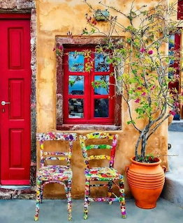 Colorful Mediterranean courtyard with red door, painted window, decorated chairs, and teracotta pot with small tree