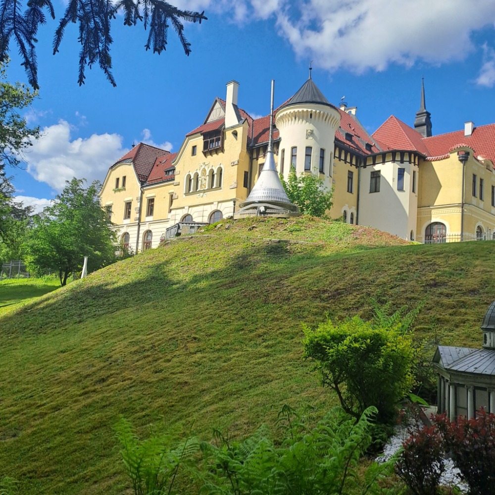 Pale yellow castle-like building with red roofs on a grassy hill beneath a blue sky