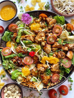 Vibrant pasta salad with chicken, mango, greens, tomatoes, and purple flowers on a patterned plate
