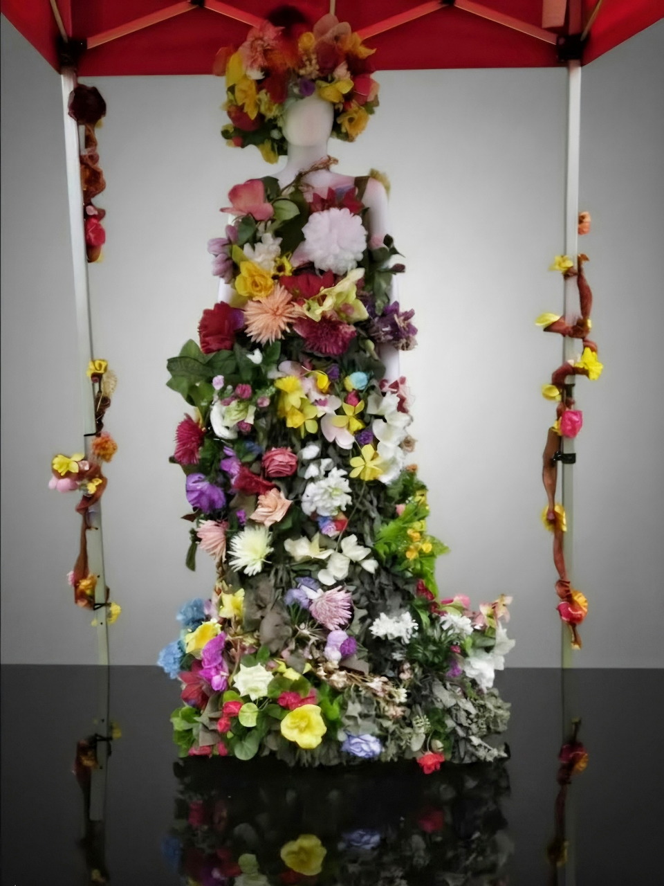 Cone-shaped sculpture covered in multicolored flowers displayed under red canopy with floral decorations