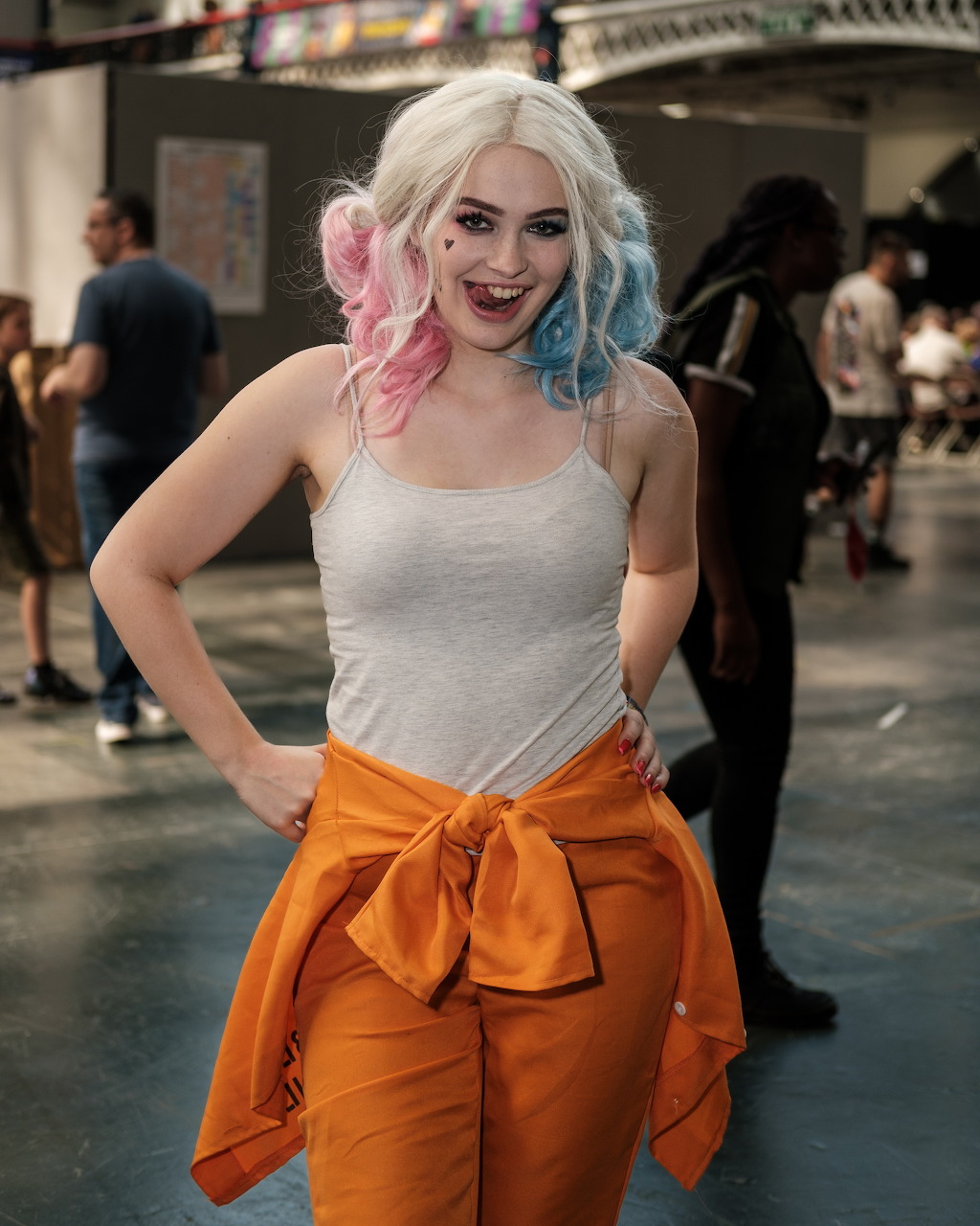 Woman cosplaying as Harley Quinn with pink and blue hair, white tank top, and orange outfit at a convention