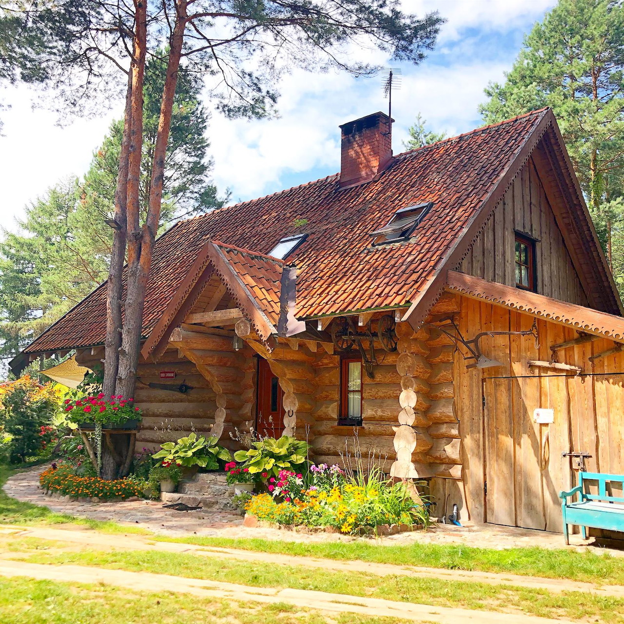 Rustic log cabin with red tile roof surrounded by pine trees and flower garden
