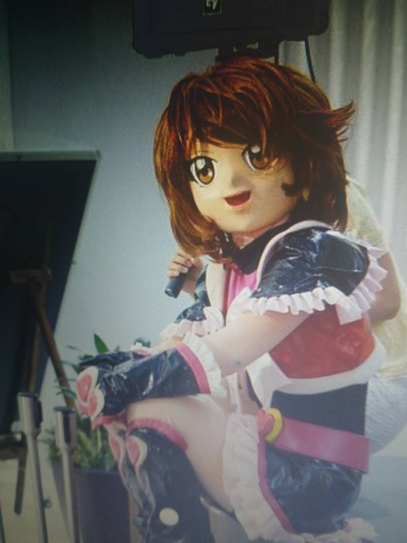 Person in anime-style kigurumi cosplay with a large character mask and pink-black outfit indoors