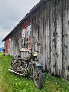 Vintage motorcycle leaning against weathered gray wooden barn with red-trimmed windows