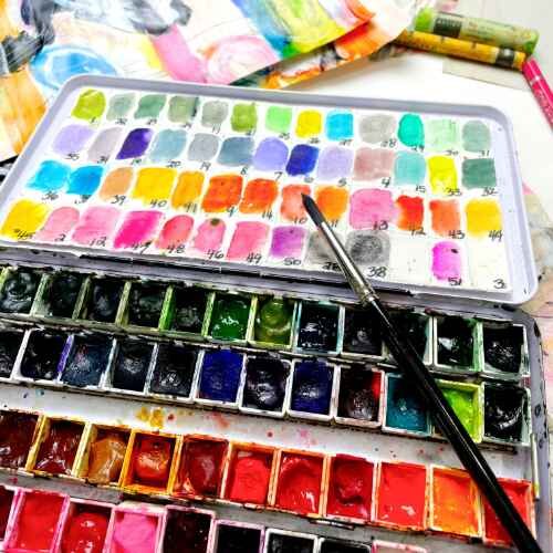 Open watercolor palette with many colorful paint wells and paintbrush resting on top