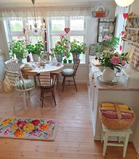 Bright vintage kitchen with wooden floors, round table, fresh flowers, poted plants, and colorful floral rug