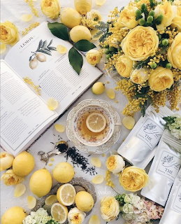 Flat lay with yellow roses, lemons, a cup of lemon tea, an open book, and tea packets on a white surface