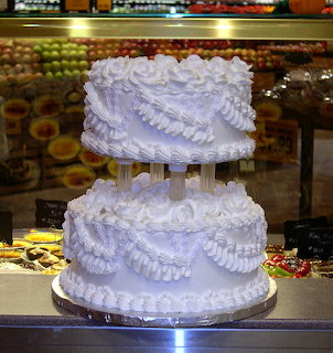 Two-tier purple wedding cake with ruffled frosting on pillared stand in bakery display