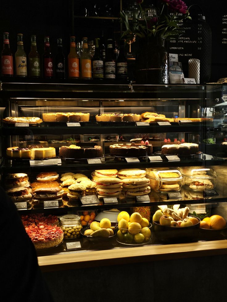 Illuminated cafe display case with cakes, pastries, and fruit on multiple shelves, liquor bottles above