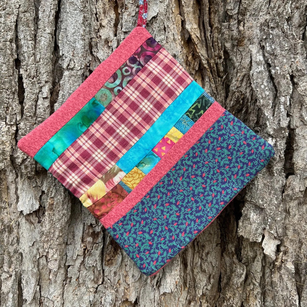 Two colorful quilted fabric pouches hanging on rough tree bark