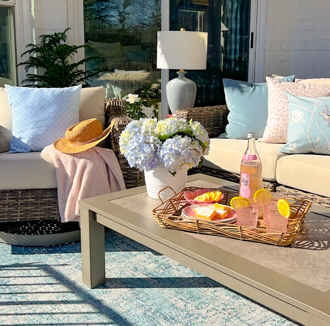 Outdoor patio seating area with white couch, blue cushions, coffee table, hydrangeas, and refreshments in sunlight