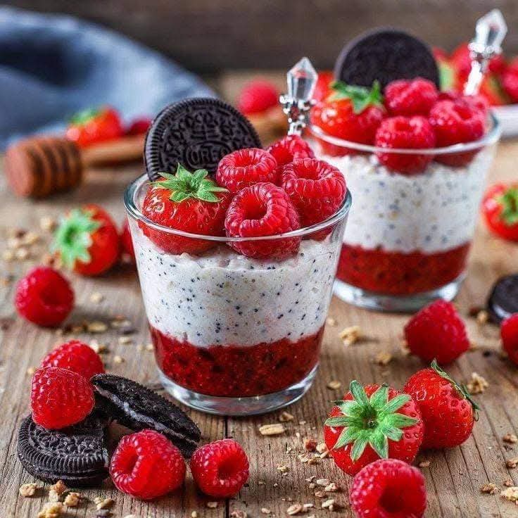 Two layered dessert cups with cream, berries, and Oreo cookies on wooden surface with fresh strawberries and raspberries