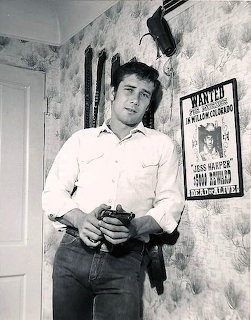Black-and-white photo of a young man with a gun standing beside a wanted poster on a wall