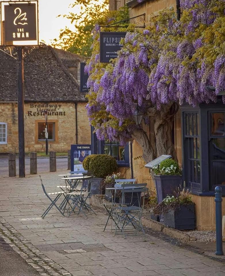 Cotswolds village street with stone buildings, cafe tables, and purple wisteria on a storefront