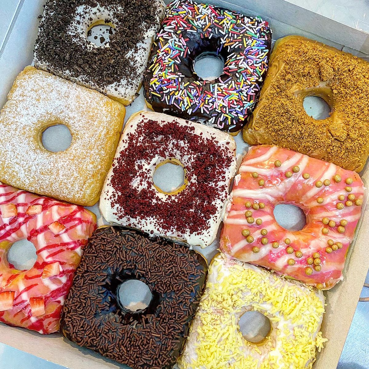 Eight square donuts with assorted toppings including chocolate, sprinkles, powdered sugar, and coconut in a white box