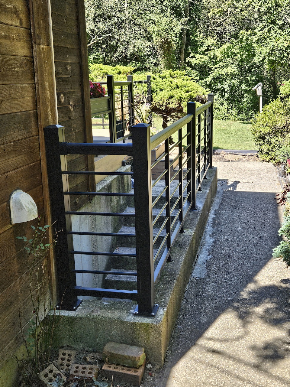 Dark aluminum horizontal railing along outdoor steps next to a wooden building with green garden in background