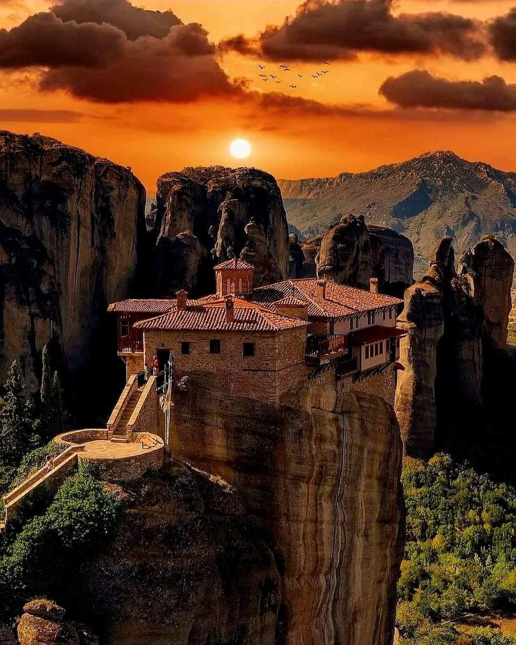 Historic monastery sitting atop sheer rock cliffs at Meteora, Greece, under a glowing orange sunset