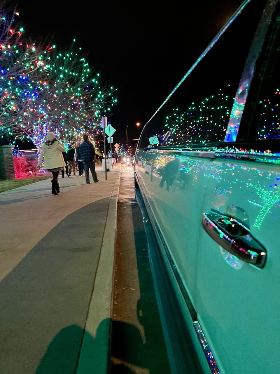 Shiny car door reflecting Christmas lights with people walking along illuminated pathway at night