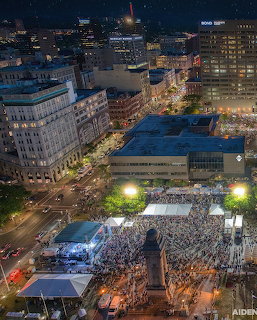 Nighttime city view of downtown Syracuse with a large crowd in a plaza and surrounding buildings