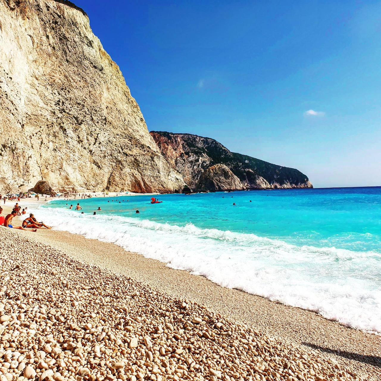 Pebble beach with turquoise sea and a tall rocky cliff beneath a clear blue sky