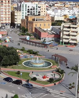 Aerial view of a city plaza with a circular fountain, paths, and nearby buildings