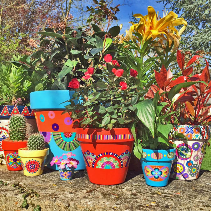Colorful painted pots with flowers, leafy plants, and cacti on a stone ledge outdoors